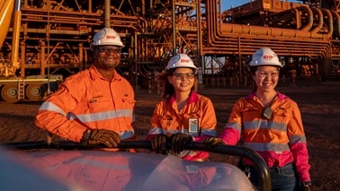 Three people in BHP orange shirts and hard hats outside processing facility